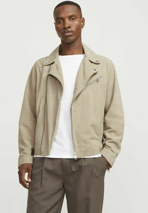 Chaqueta de biker de ante beige con cremallera asimétrica, cuello de presión y bolsillos laterales, usada sobre una camisa blanca y pantalones marrones.