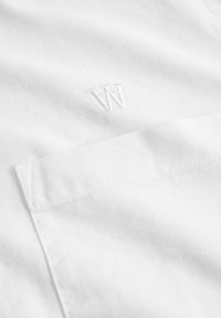 Tela de algodón blanca con una textura suave, que presenta un sutil logo bordado en la esquina inferior y un bolsillo de parche en el lado izquierdo.