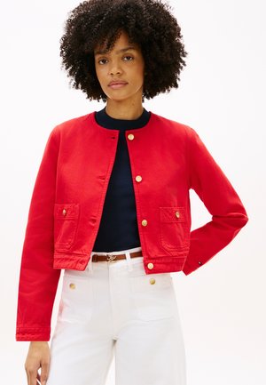 Rote, kurz geschnittene Jacke aus Baumwolle mit rundem Ausschnitt, goldenen Knöpfen, zwei Fronttaschen und strukturierter Oberfläche. Wird über einem marineblauen Oberteil getragen.