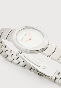 Ασημένιο ρολόι χειρός Calvin Klein με μινιμαλιστικό λευκό καντράν, ροζ δείκτες και μπρασελέ από ανοξείδωτο ατσάλι με κούμπωμα.