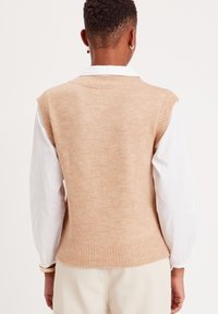 Chaleco de punto beige con dobladillo y sisas acanalados, llevado sobre una camisa blanca con cuello. Textura suave y ajuste relajado.