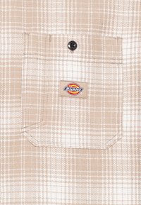 Beżowa i biała tkanina w kratę z kieszenią zapinaną na guzik, posiadającą czarny guzik oraz prostokątną naszywkę z logo Dickies.