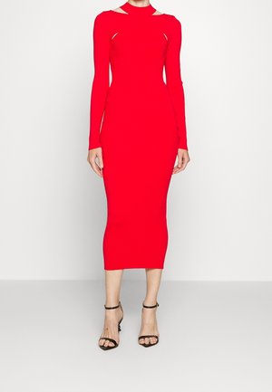 Kobieta w obcisłej, długorękawowej, czerwonej sukience midi z wycięciami na ramionach i czarnych sandałach na wysokim obcasie stojąca na jednolitym tle.