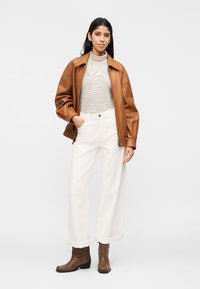 Γυναίκα που στέκεται με το ένα χέρι στην τσέπη, φοράει καφέ δερμάτινο μπουφάν, ριγέ ζιβάγκο, λευκό παντελόνι με φαρδιά πόδια και καφέ μποτάκια μέχρι τον αστράγαλο.