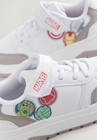Zapatillas blancas con acentos grises, con gráficos de personajes de Marvel que incluyen a Iron Man y Hulk. Diseño con tira de Velcro y cordones.
