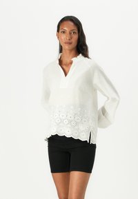 Hvit brodert bluse med V-hals, volangkrage og bølgekant. Har blomsterdesign og lett, gjennomsiktig materiale. Sorte shorts.