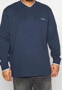 Marineblauwe polo met lange mouwen, voorzien van een knoopjeslijst en witte kraagafwerking. Heeft een klein Calvin Klein-logo op de linkerkant van de borst. Gemaakt van een katoenmix.