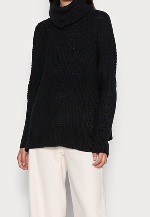 Suéter de cuello alto de punto grueso negro con mangas largas, combinado con pantalones blancos de corte holgado en una persona de pie.