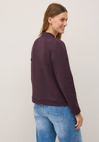 Woman with shoulder-length hair, wearing a purple sweatshirt and blue jeans, smiles gently while looking over her shoulder against a neutral background.