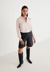 Helles rosa Bluse mit V-Ausschnitt und gerafften Ärmeln, kombiniert mit dunkel gestreiften Shorts. Schwarze knielange Stiefel runden das Outfit ab.