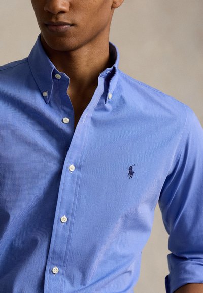 Bărbat purtând o cămașă albastru deschis cu nasturi, având un mic logo brodat închis reprezentând un jucător de polo în partea stângă a pieptului.