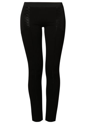 Femme portant un sweat-shirt gris avec un grand visage de dessin animé noir et blanc, un pantalon noir ajusté, des chaussures à talons noires, et tenant un petit sac à main blanc.