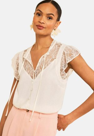 Žena nosi biely tielkový top s čipkovými detailmi a šnúrkami, v kombinácii s svetloružovými nohavicami s vysokým pásom, pózuje s rukou na boku.