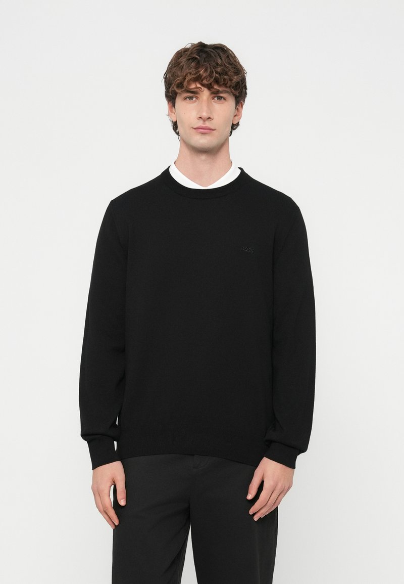 Czarny sweter z gładkiego dzianinowego materiału, z okrągłym dekoltem i długimi rękawami. Dyskretne logo na piersi, zestawiony z ciemnymi spodniami.