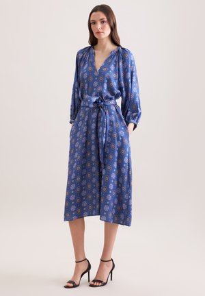 Kobieta ubrana w niebieską sukienkę midi w wzory z długimi rękawami i paskiem zawiązanym w talii, w połączeniu z czarnymi sandałami na wysokim obcasie.