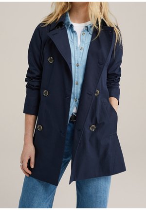 Kobieta ubrana w granatowy, dwurzędowy płaszcz na jasnoniebieską koszulę dżinsową i niebieskie jeansy, z jedną ręką w kieszeni płaszcza, a drugą opuszczoną wzdłuż ciała.