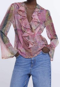 Blusa rosa trasparente con scollatura arricciata e maniche a sbuffo. Presenta motivi colorati e intricati sul tessuto. Indossata con jeans blu.