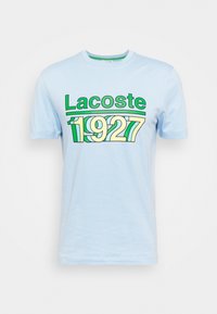 Camiseta de algodón azul claro con gran estampado en verde y amarillo "Lacoste 1927" en el frente. Cuello redondo clásico y mangas cortas.