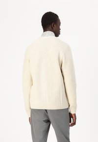 Suéter de punto color crema con un patrón texturizado, mangas raglán y puños acanalados. Combinado con pantalones grises.