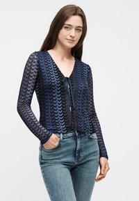 Γυναίκα με μακριά καστανά μαλλιά, φορώντας μπλε μπλούζα με σχέδια και μακριά μανίκια με μπροστινό δέσιμο, συνοδευόμενη από μπλε τζιν τζιν, που στέκεται με το ένα χέρι στην τσέπη.