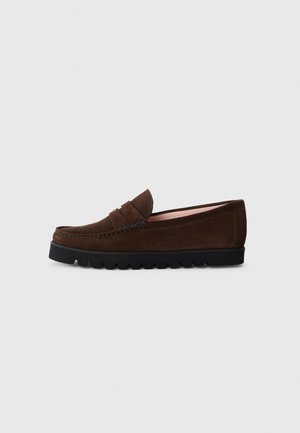Mocassin en daim brun foncé avec une semelle en caoutchouc noire et une lanière décorative, présenté de côté sur un fond gris clair.
