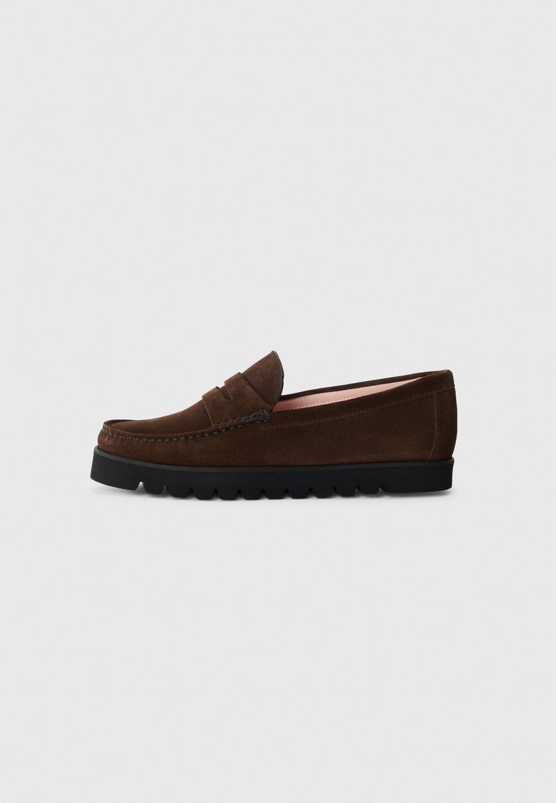 Mocassin en daim brun foncé avec une semelle en caoutchouc noire et une lanière décorative, présenté de côté sur un fond gris clair.