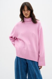 Maglione lavorato a maglia oversize di colore rosa, con collo alto, orlo a coste e spalle scese, abbinato a pantaloni di jeans blu larghi.