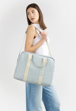 Donna con top bianco senza maniche e jeans blu che porta una grande borsa a tracolla grigio chiaro con manici e tracolla beige.