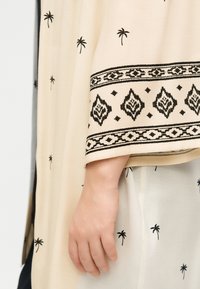 Χέρι ακουμπισμένο σε μπεζ ύφασμα με μαύρα μοτίβα φοίνικα και γεωμετρικά περιγράμματα στο μανίκι και το παντελόνι.