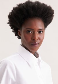 White button-up shirt with a structured collar, paired with dark, textured curly hair. The background is neutral, highlighting the shirt.