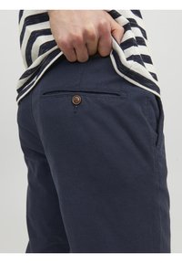 Mörkblå byxor i en texturerad tyg, med en bakficka med en brun knappdetalj, ihop med en randig skjorta.