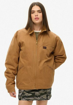 Chaqueta marrón con cremallera y cuello alzado, que cuenta con bolsillos laterales y un pequeño logotipo. Textura suave y corte holgado.