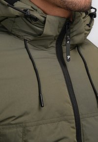 Geacă izolată de culoare verde măslinie, cu fermoar în față, șnururi ajustabile și accente negre la fermoar. Material texturat și detalii la glugă vizibile.
