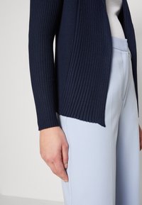 Námořnický žebrovaný kardigan s otevřeným předním dílem a dlouhými rukávy, spárovaný se světle modrými kalhotami na míru, který vykazuje hladké textury a přiléhavý střih.