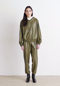 Ensemble de survêtement en simili cuir verni vert olive avec capuche, taille élastique et poignets. Associé à des bottines noires à la cheville. Design simple et moderne.
