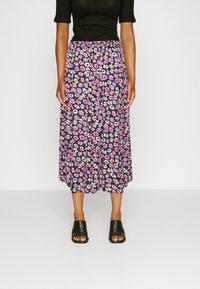 Femme portant un haut noir côtelé, une jupe mi-longue fleurie avec des fleurs roses et violettes, et des sandales mules noires à bouts ouverts, debout sur un fond uni.