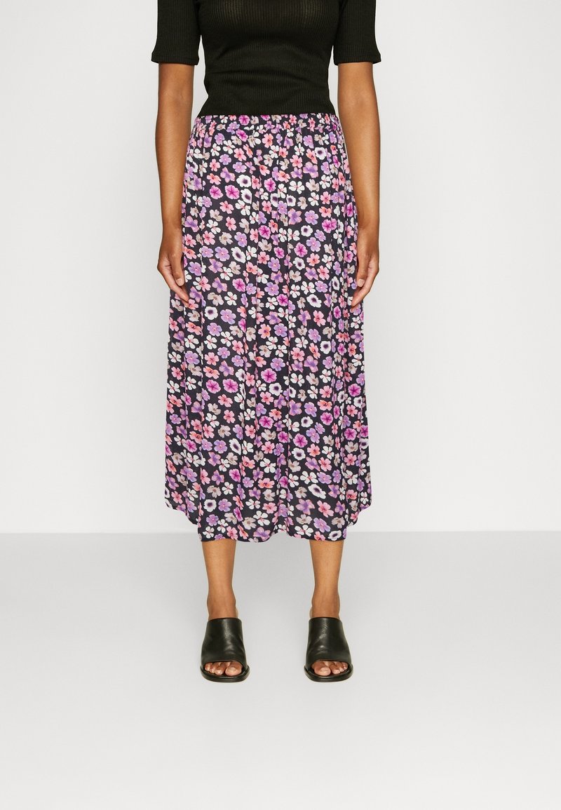 Femme portant un haut noir côtelé, une jupe mi-longue fleurie avec des fleurs roses et violettes, et des sandales mules noires à bouts ouverts, debout sur un fond uni.