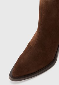 Bottine en daim marron avec un bout pointu, texture lisse et un détail de couture discret ; présente un talon et une semelle solides de couleur foncée.
