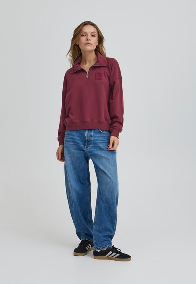 Burgundska zip-sweatshirt s visokim ovratnikom i izvezenim logotipom, uparen s labavim plavim trapericama i crnim tenisicama.