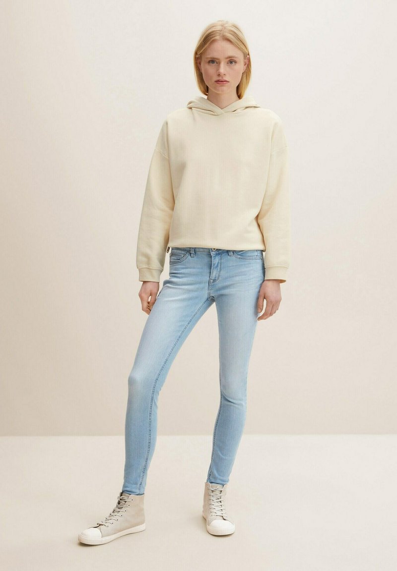 Krämfärgad hoodie med en avslappnad passform och känguruficka, i kombination med ljusblå tighta jeans och grå hög-top sneakers.