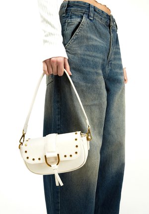 Osoba ubrana w niebieskie dżinsy i biały sweter trzymająca małą białą torebkę z złotymi ćwiekami i klamrą.