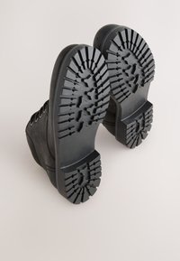 Schwarze Knöchelstiefel mit einer strukturierten Gummisohle. Verfügt über ein Profil mit tiefen Stollen für besseren Halt und Metallösen für die Schnürsenkel. Obermaterial aus glattem Veloursleder.