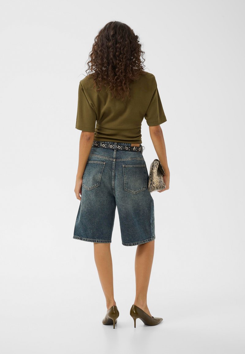 Femme aux cheveux bouclés, vue de dos, portant un haut à manches courtes vert olive, un short en jean jusqu’aux genoux, des talons et tenant une pochette en cuir de serpent.