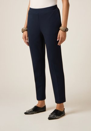 Pantaloni navy a lunghezza caviglia in tessuto liscio con una vestibilità su misura, abbinati a scarpe slip-on nere con accenti di borchie argentate.