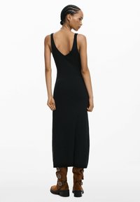 Nő fonott hajjal, fekete ujjatlan midi ruhában és barna, csatos bokacsizmában, háttal a fehér háttér előtt áll.