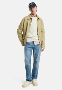 Beige bomuldsjakke over en hvid kabelstrikket sweater, lyseblå jeans og cremede sneakers. Mørkeblå kasket med hvid tekst fuldender looket.