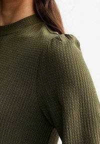 Primo piano di una persona che indossa un top in tessuto verde oliva con una scollatura alta e dettagli con spalle a sbuffo.