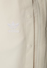 Krämfärgad skjorta med texturerat tyg, knäppning och en vit broderad Adidas-logotyp med tre lövdekorationer.