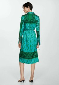 Rochie verde cu un model din material ușor, având mâneci lungi, un cordon la talie și guler. Designuri geometrice notabile în nuanțe variate.