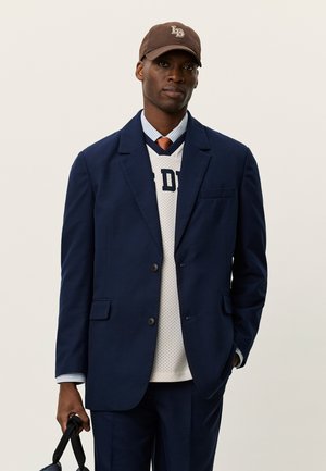 Marineblå dressjakke med to knapper over en hvit mesh-genser. Brun caps med beige logo. Lyseblå stripete skjorte og rustfarget slips.